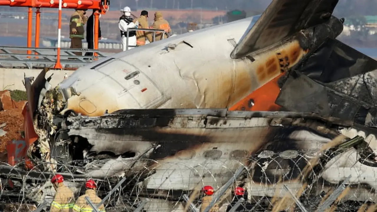 Workers in uniforms inspect the burned fuselage of a jetliner, which is lying on an airport runway.